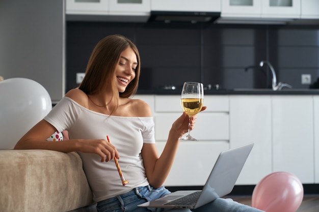 Smiling young woman with glass of white wine sitting at home and having video call on modern laptop