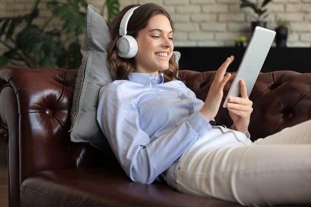 Smiling young woman with headphones and digital tablet on the sofa.