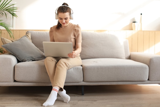 Smiling young woman with headphones and laptop on the sofa.