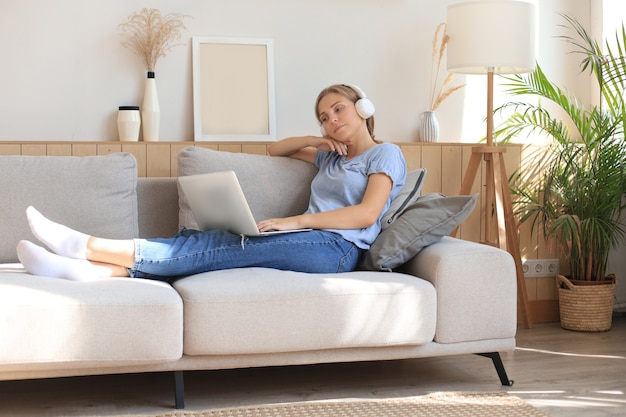 Smiling young woman with headphones and laptop on the sofa.