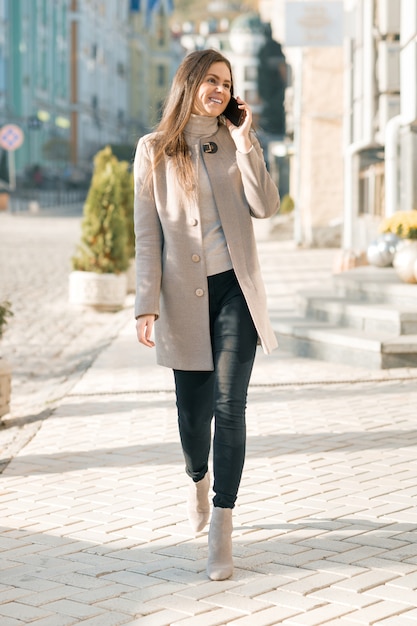 Smiling young woman with mobile phone walking in the sunny city street,