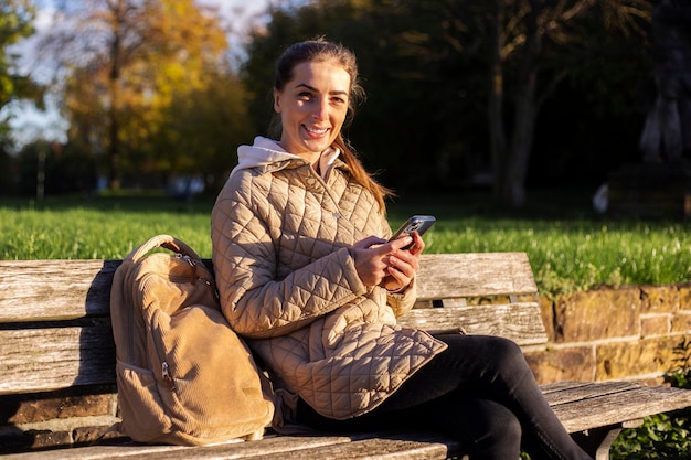 Smiling young woman with a phone sitting on a bench in the park