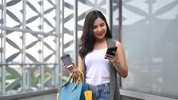 Smiling young woman with shopping bags using smartphone and walking outside of shopping mall