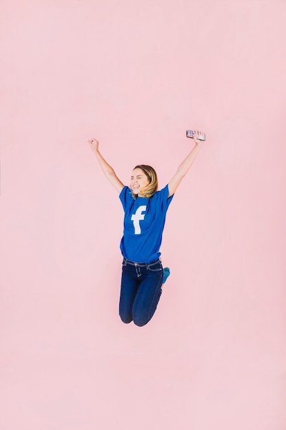 Smiling young woman with smartphone jumping on pink background