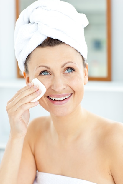 Smiling young woman with a towel putting cream on her face in the bathroom