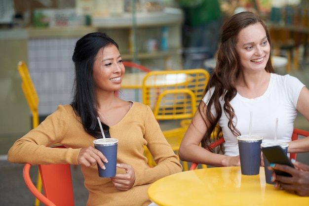 Smiling young women sitting at cafe table drinking iced tea and listening to friends telling story