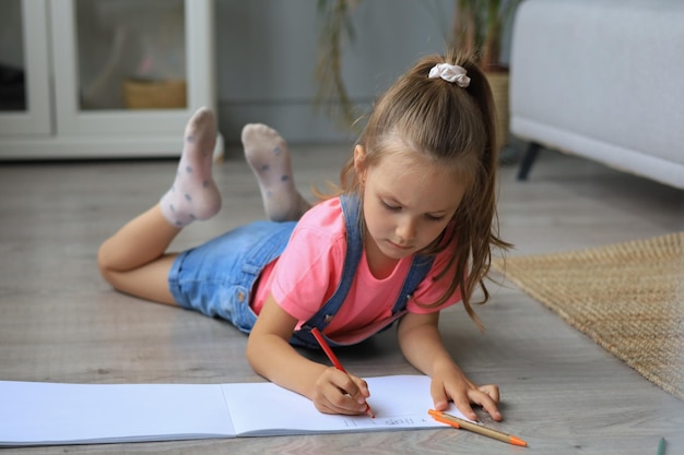 Smilling happy girl lying on warm floor with a toy elephant near to her enjoying creative activity drawing pencils coloring pictures in albums