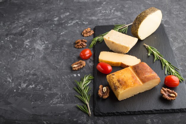 Photo smoked cheese and various types of cheese with rosemary and tomatoes on black slate board on a black concrete surface