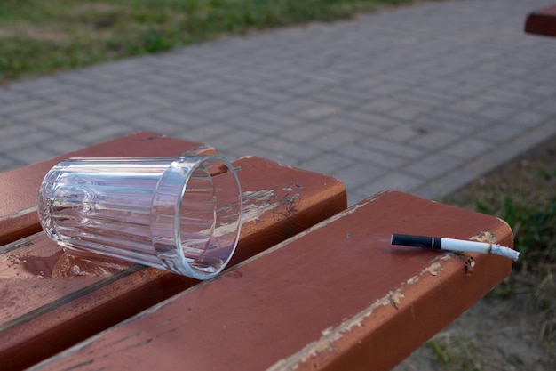 Smoking cigarette and emptied glass on brown bench on background of green grass and gray sidewalk