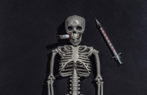 Smoking and drug addiction. Smoking skeleton with cigarette in the mouth and syringe on black dark background. Smoking and drugs kills