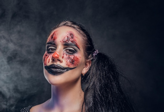 In smoky dark photo studio woman in a role of evil clown is posing for photographer.