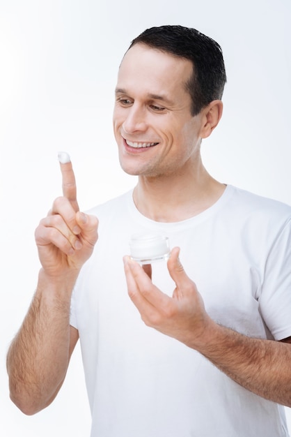 Smooth skin. Positive nice handsome man holding a bottle of cream and looking at his index finger while applying the cream on his face