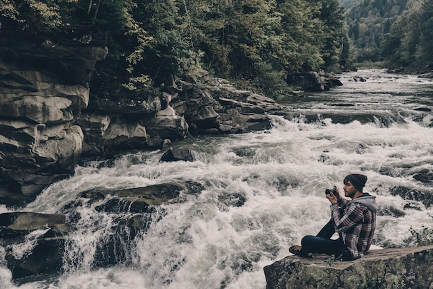 Snapping memories. Handsome young modern man photographing while sitting on the rock near the river