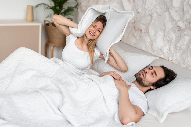 Snoring young guy and his frustrated girlfriend covering ears with pillow in bed at home