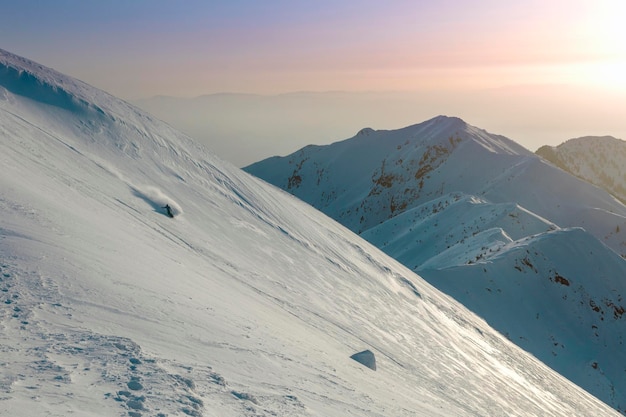 A snowboarder descends a wide slope against the background of mountain ranges and the sunset