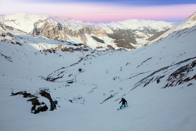 Snowboarder on a winter ski resort in front of mountains covered with snow under a gentle pink sky