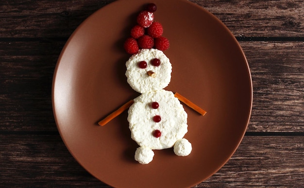 Snowman made of cottage cheese and raspberries and lingonberries on a brown plate on a wooden background