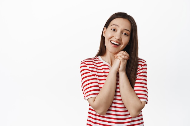 So adorable. Cute brunette girl sighing, looking at something lovely and touched, clench hands and smiling, gazing and admire smth, standing against white background