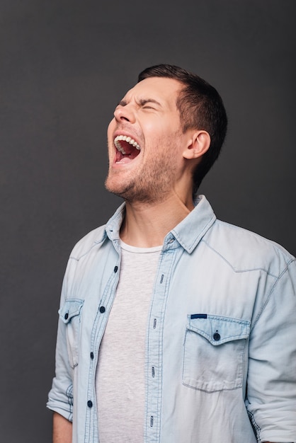 So furious! Young handsome man keeping mouth open and eyes closed while standing against grey background