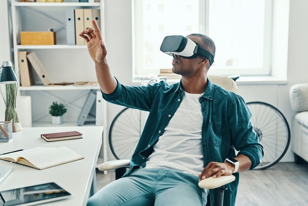 So real. Handsome young African man wearing virtual reality simulator headset while sitting indoors