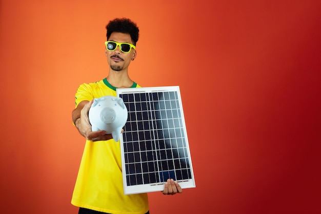 Soccer Player Black Man Celebrating Holding Solar Photovoltaic Panel and Pig Coin Isolated on Orange Background