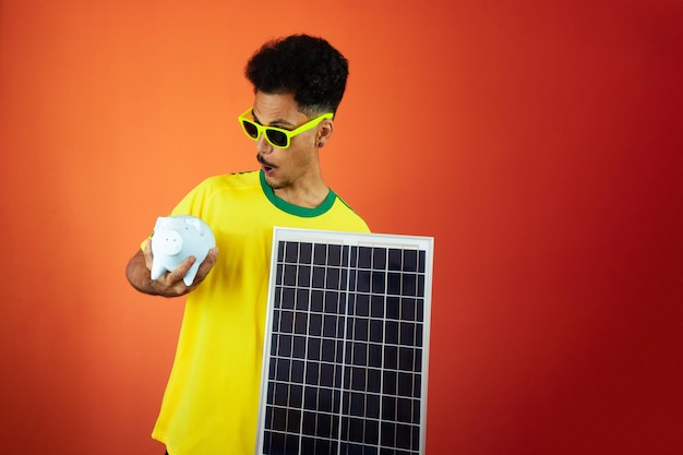 Soccer Player Black Man Celebrating Holding Solar Photovoltaic Panel and Pig Coin Isolated on Orange Background