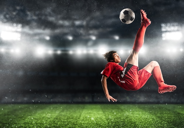 Soccer striker in red uniform hits the ball with an acrobatic kick in the air at the stadium