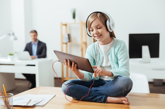 Sociable lively diligent child using her gadget while wearing headphones and sitting on the table in dads office