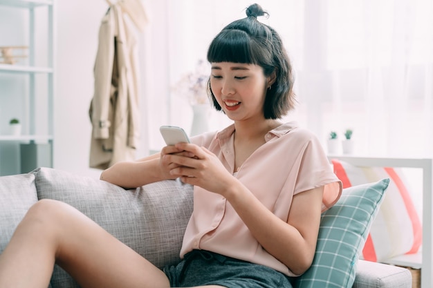 Social networks concept. Young asian japanese woman messaging on smartphone at home living room sitting comfortably on sofa. cute cheerful girl using cellphone relax on couch in cozy apartment