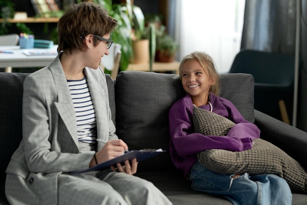 Social worker making notes and having positive conversation with little girl while they sitting on s