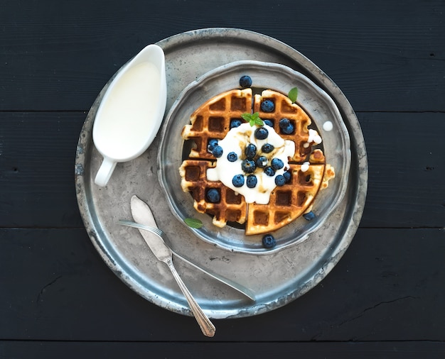 Soft Belgian waffles with blueberries, honey and whipped cream on vintage metal plate over black wooden table, top view.