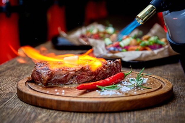 Soft focus. Entrecote Beef Grilled steak meat with fire flames on wooden cutting boardon with rosemary branch, pepper and salt . Master chef cooking delicious grill barbecue. Melt butter