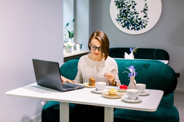 Soft focus shot of woman at coworking space or cafe with internet connection, work on laptop and enjoy desserts, waiting for clients. Concentrated at work.