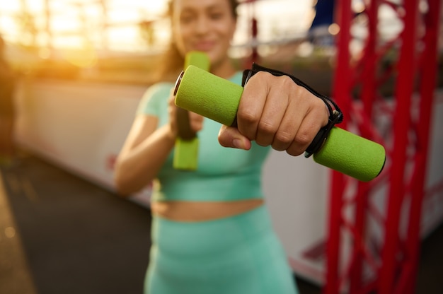 Photo soft focus on sporty woman's outstretched hand holding a small dumbbell, standing on the background of a boxing arena. blurred fitness woman exercises outdoors at sunrise