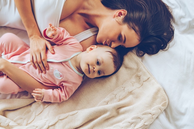 Soft kiss from mommy. Top view of cheerful beautiful young woman lying in bed with her baby girl and kissing her