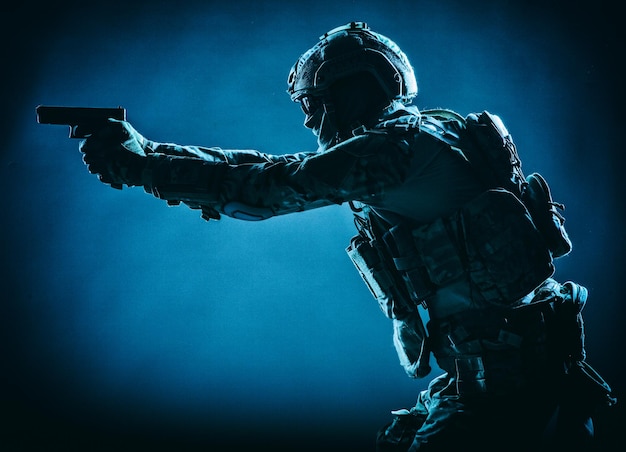 Soldier of army elite forces, special security service fighter with hidden behind mask and glasses face, in helmet and load carriage system, aiming with service pistol low key, side view, studio shoot