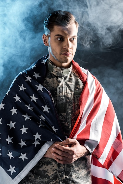 Soldier in military uniform with clenched hands and american flag on black with smoke