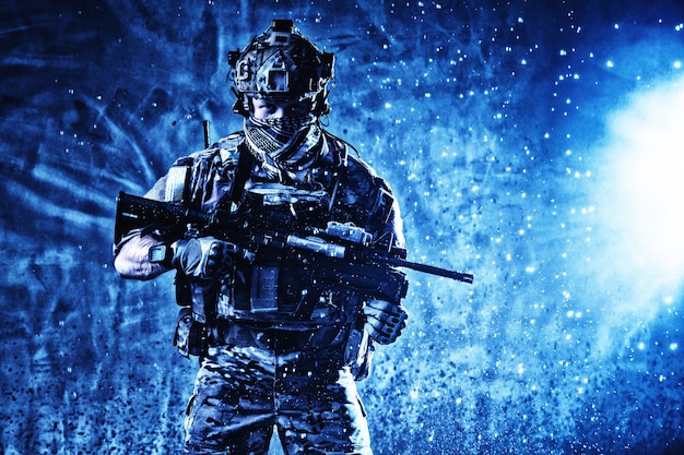 Soldier in tactical ammunition, wearing camo uniform, battle helmet, radio headset, hiding face behind scarf, standing with assault rifle in sparkles splash from explosion beside, looking in camera