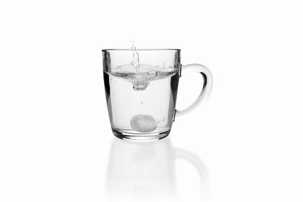 Soluble fizzy tablet fallen with splash to transparent glass mug on white surface.