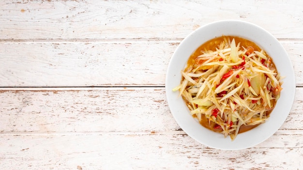 Som Tum Pla Ra Thai ESan food spicy papaya salad with pickled fish tomato lime and chilli in white ceramic plate on white wood texture background with copy space for text top view