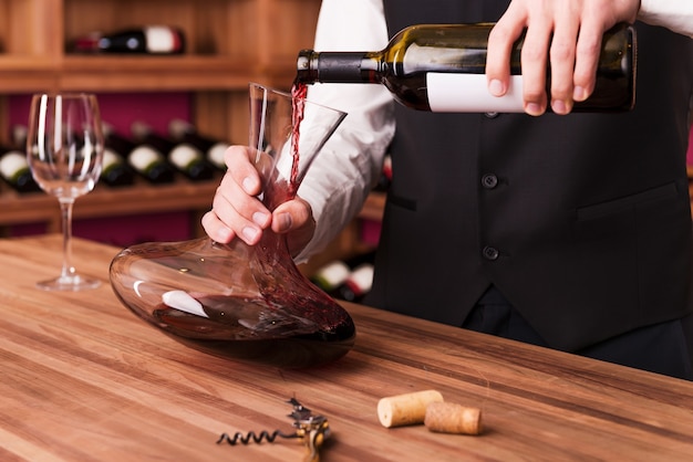 Sommelier at work. Confident male sommelier pouring wine to decanter while standing near the wine shelf