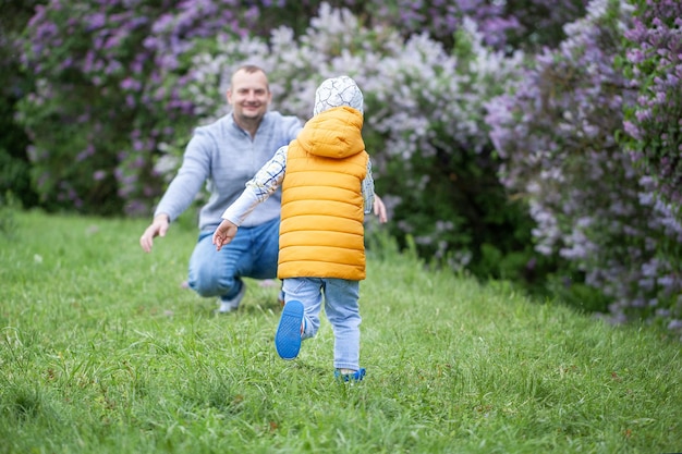 Son runns into father's arms. Man crouching at the park with his arms outstretched, with a boy running towards him. Back view.