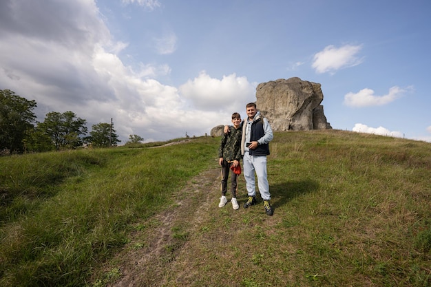 Son with father wear backpack hiking near big stone in hill Pidkamin Ukraine