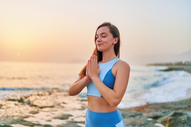 Soncentrated blissful one fitness woman with closed eyes and praying hands standing alone with meditation position while breathing exercises by sea Mental mind care and healthy habits