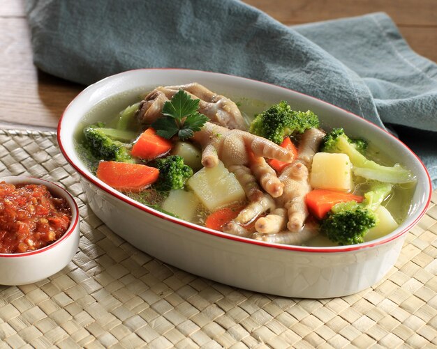 SOp Ceker Ayam or Clear Chicken Feet (Claw) Soup with Potato, Broccoli, and Carrots. Served on Marble Table in Brown Bowl with Sambal