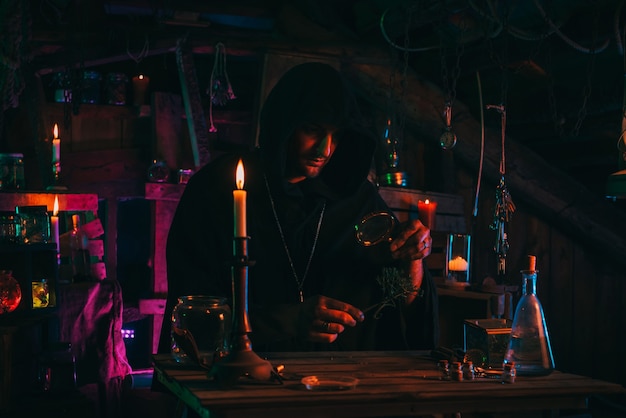 Sorcerer alchemist in dark clothes is engaged in potion making in a craft workshop with a colorful neon light