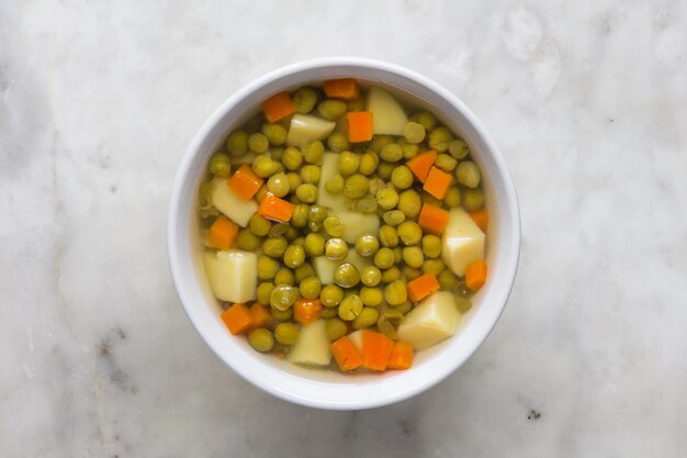 Soup with green peas and vegetables in white bowl on marble background