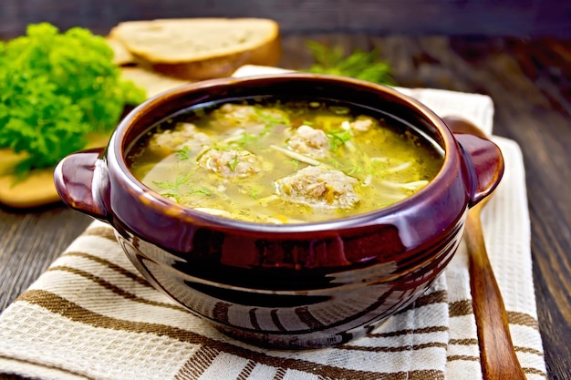 Soup with meatballs noodles and vegetables in a clay bowl spoon on a napkin parsley bread on a wooden boards background