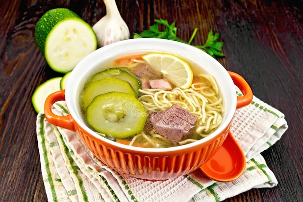 Soup with zucchini beef ham lemon and noodles in a bowl parsley and dill on a towel on a wooden board background