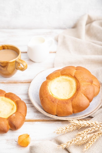 Sour cream bun with cup of coffee on a white wooden background and linen textile. Side view, close up, selective focus.